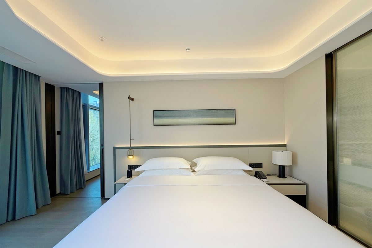 A modern hotel room with a large bed, bedside tables, a nightstand, a wall-mounted lamp, and a window with curtains.