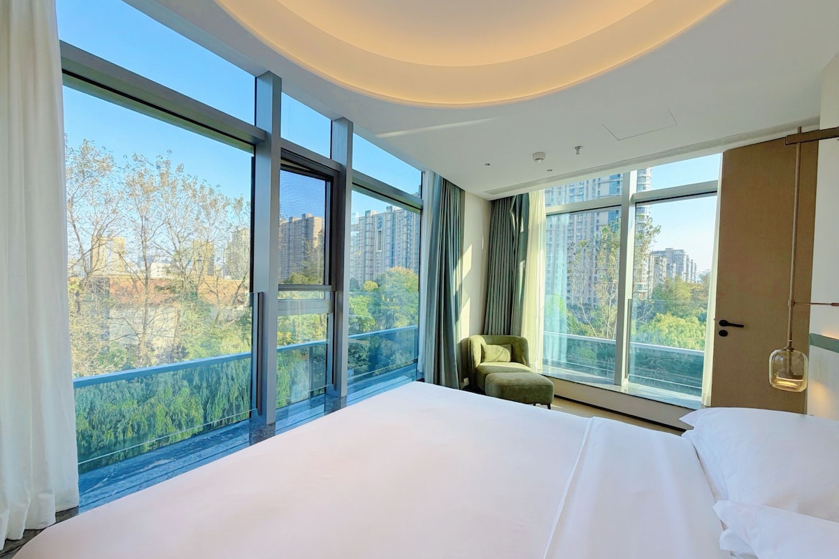 A hotel room with a large bed, a chair, and a view of the cityscape through large windows.