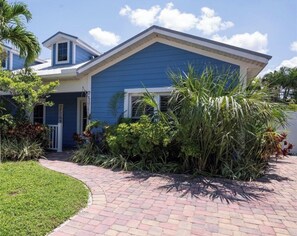 Uma casa de dois andares com fachada azul, um caminho de tijolos até a entrada e vegetação tropical ao redor da propriedade.