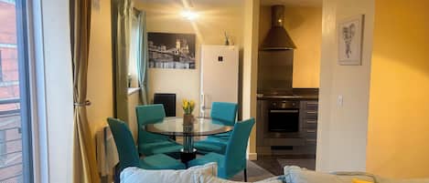 A modern living room with teal and yellow cushions, a round glass table with a vase, and a kitchen with a stainless steel range hood.