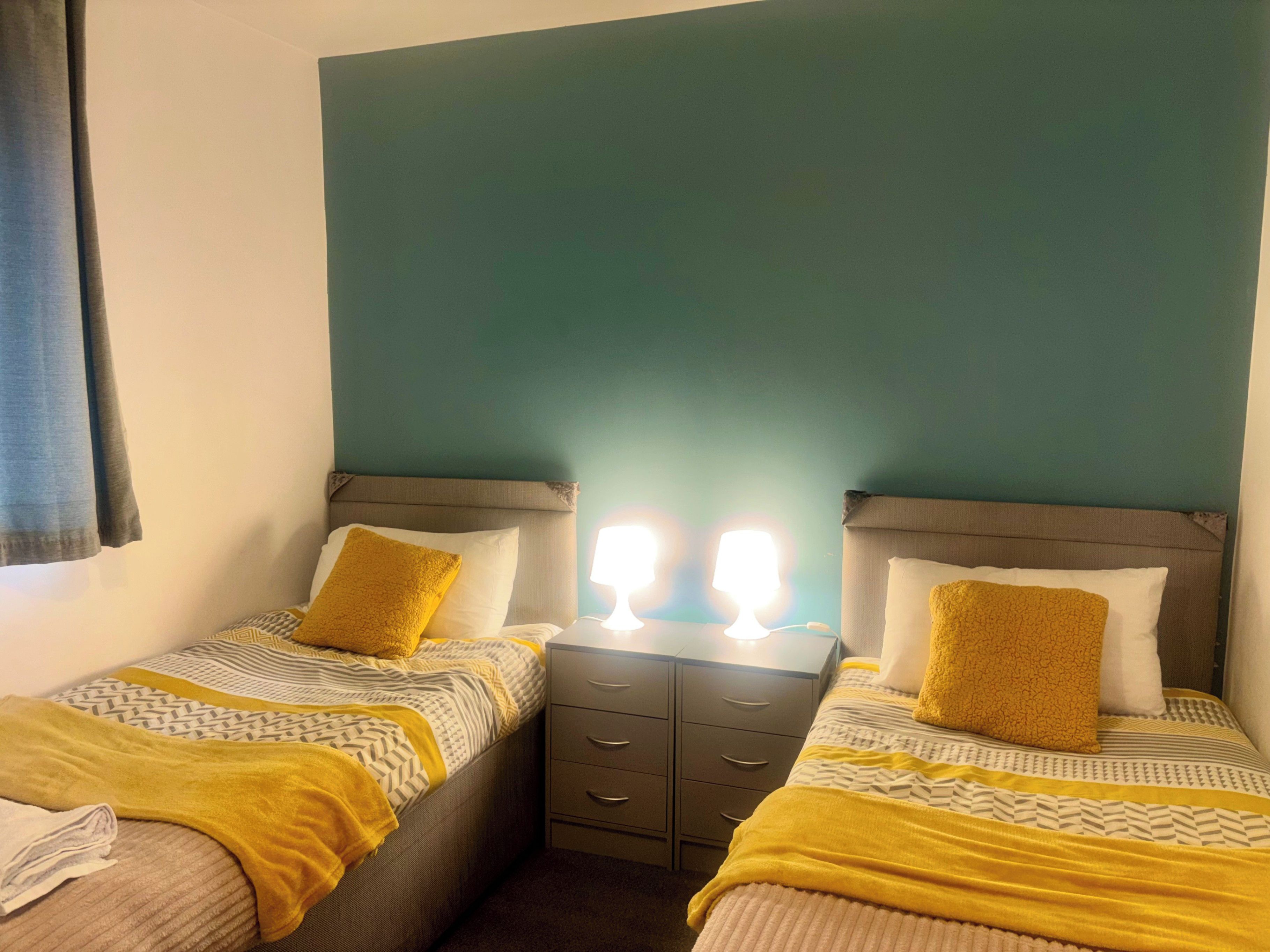 Quarto duplo com duas camas, uma mesa de cabeceira e uma luminária fixada na parede.
