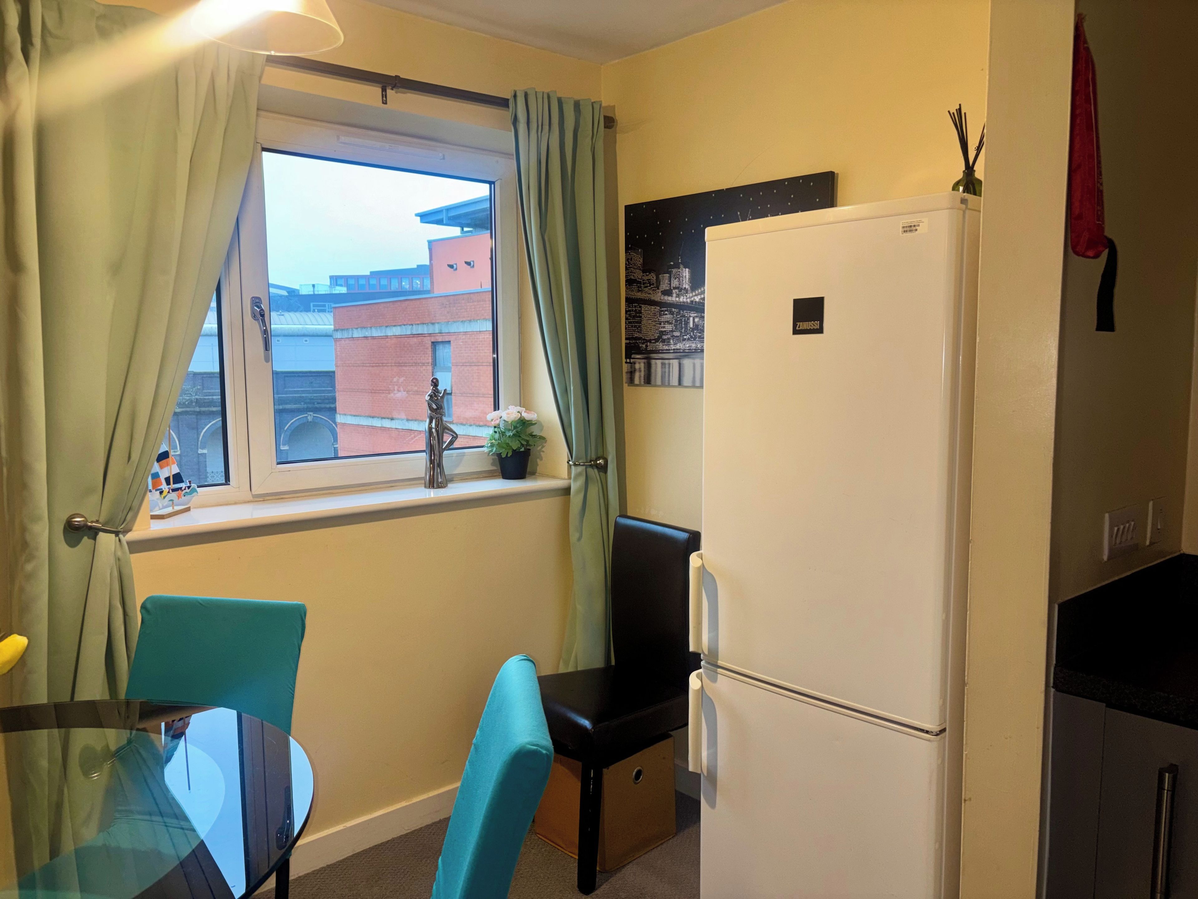 A compact kitchen with a refrigerator, a window with curtains, a small table with chairs, and a potted plant on the windowsill.