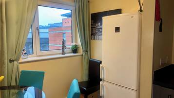 A compact kitchen with a refrigerator, a window with curtains, a small table with chairs, and a potted plant on the windowsill.