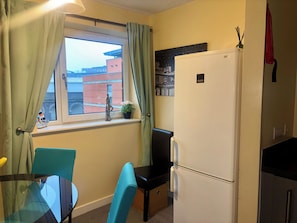 Cozinha compacta com geladeira, janela com cortinas, mesa pequena com cadeiras e uma planta em vaso no parapeito.