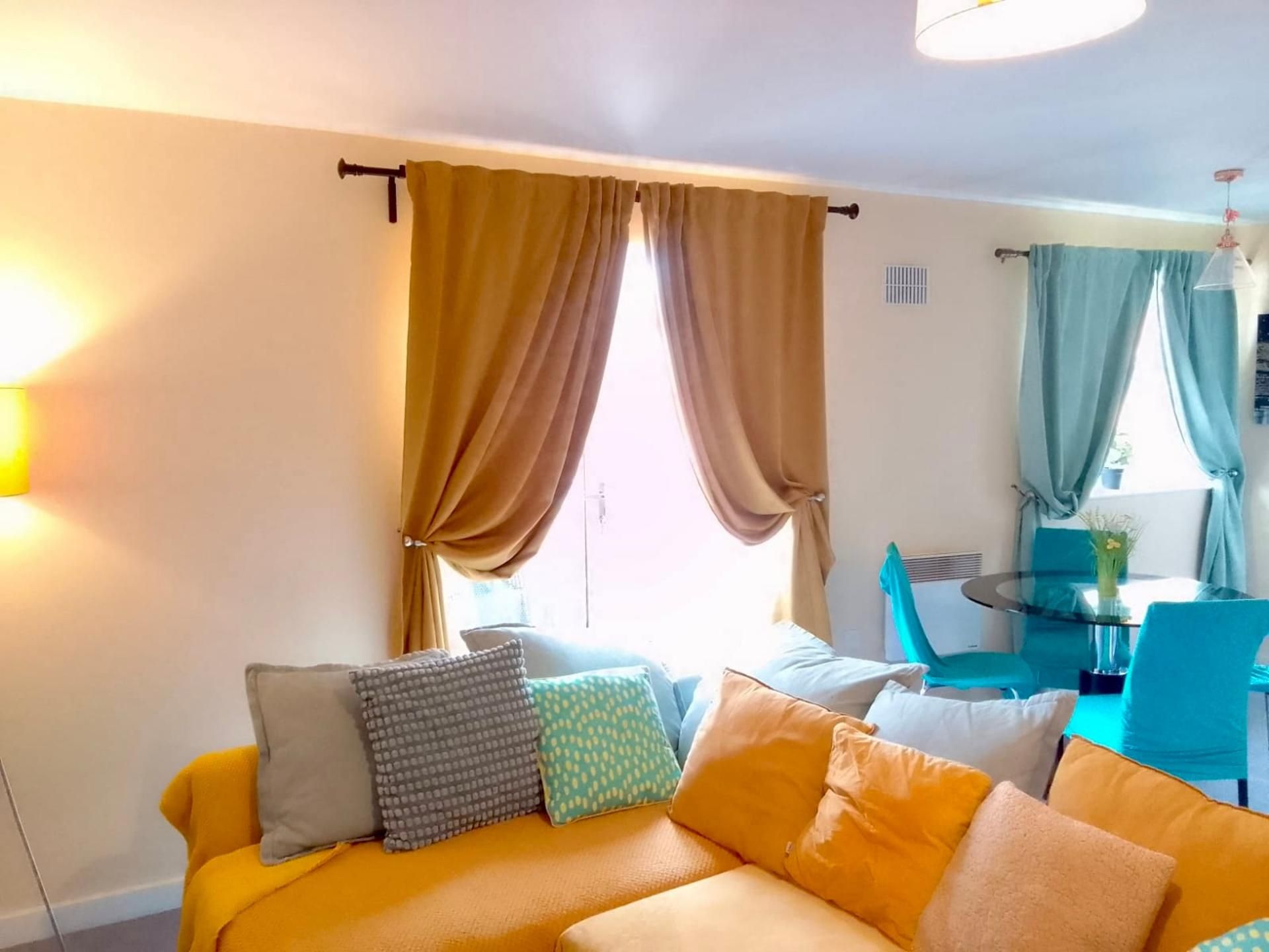 A room with a yellow sofa, multiple pillows, a dining table with chairs, and a window with curtains.