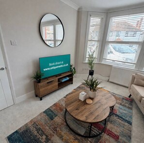 Un salon avec une table basse ronde, un téléviseur posé sur un meuble en bois, un miroir accroché au mur et une vue sur l’extérieur grâce à de grandes fenêtres.