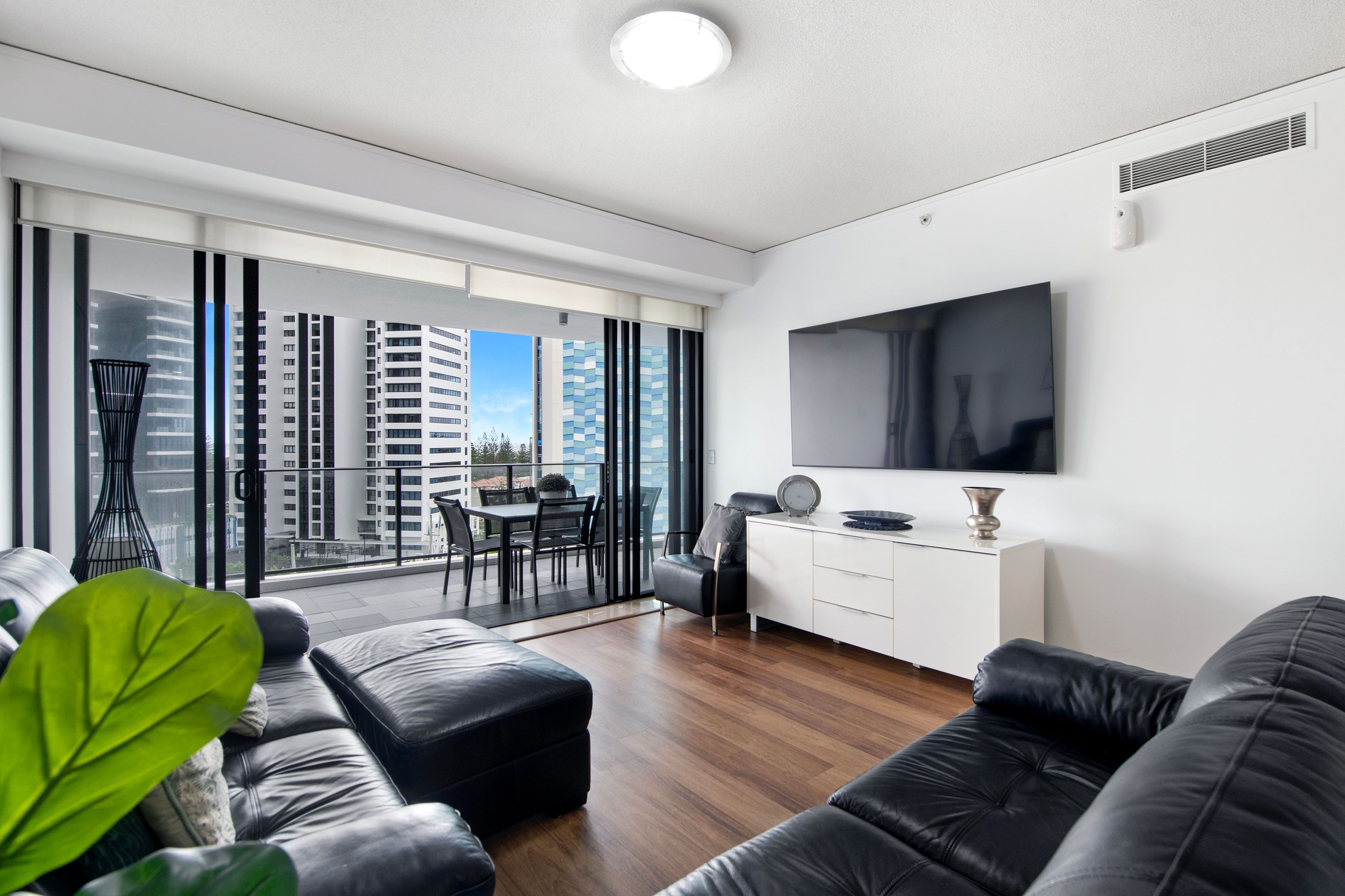 A modern living room with a sofa, a dining table with chairs, a TV mounted on the wall, and a balcony with a view of high-rise buildings.