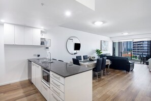 A modern kitchen with a central island, a dining area with a round table, and a view of a cityscape through large windows. - Sierra Grand Spacious 3 Bedroom Apartment (Broadbeach)