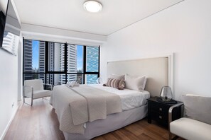 Ein modernes Schlafzimmer mit einem großen Bett, einem Nachttisch, einem Sessel und Blick auf hohe Gebäude durch das Fenster.