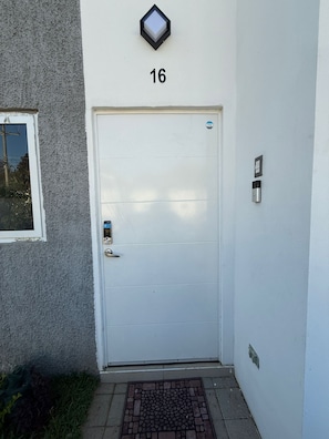 Eine weiße Tür mit der Aufschrift '16' darüber, einem kleinen Fenster links sowie einem Sprech- und Klingelsystem rechts.