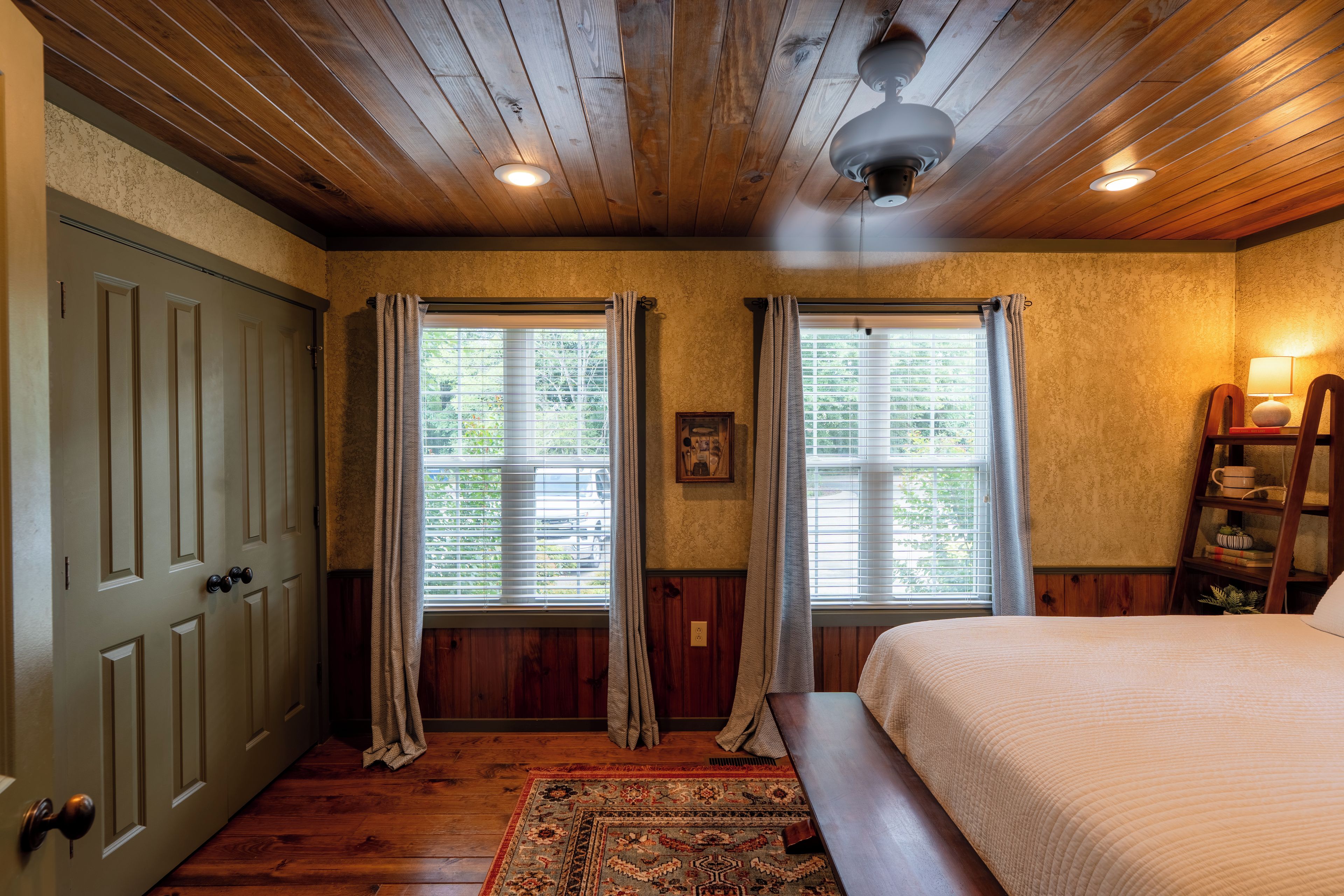 Un dormitorio con techo de madera, una cama, una mesita de noche, una lámpara, una ventana con cortinas y una puerta.
