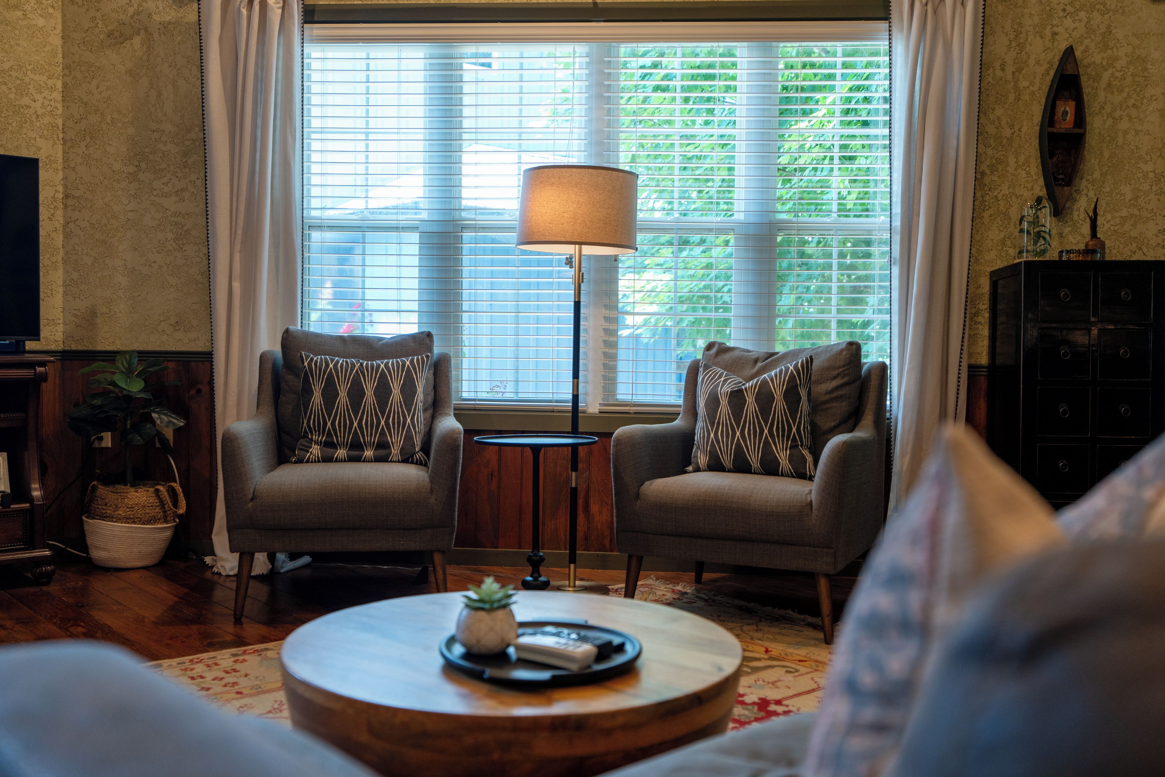 Een woonkamer met twee fauteuils, een vloerlamp, een salontafel met een plant en uitzicht door een raam met jaloezieën.