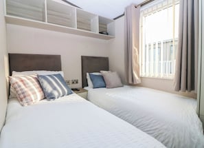 A hotel room with two beds, a headboard, a nightstand, a power outlet, and a window with curtains. - Cabin Sleeps 6 with Parking (Eyemouth)