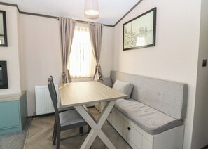 A compact room with a dining table, a sofa, a radiator, a window with curtains, and a framed picture on the wall. - Cabin Sleeps 6 with Parking (Eyemouth)
