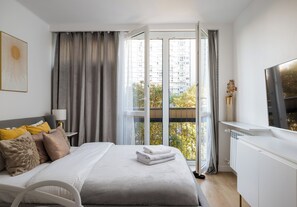A modern hotel room with a bed, a bedside table, a lamp, a window with curtains, and a view of the outside. - Park Mirowski Signature Apartment (Warszawa)