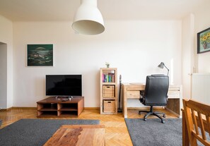 A living room with a flat-screen TV, a wooden coffee table, a bookshelf, a desk with a chair, and a hanging light fixture. - Metro Racławicka Cosy Apartment (Warszawa)