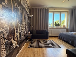 A room with a cityscape mural, a sofa, a bed, a window with curtains, and a radiator. - Dworcowa Old Town Studio (Wrocław)