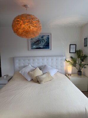 A bedroom with a large bed, a tufted headboard, a hanging orange light fixture, a framed abstract painting, and a potted plant on a bedside table.