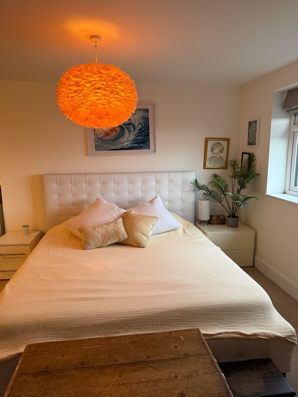 A bedroom with a large bed, a tufted headboard, a hanging orange light fixture, a framed artwork, a bedside plant, and a window with a view.