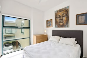 A modern bedroom with a large window, a bed with white bedding, a wooden dresser, and artwork on the walls. - Upstairs Resort Apartment (Anglesea)