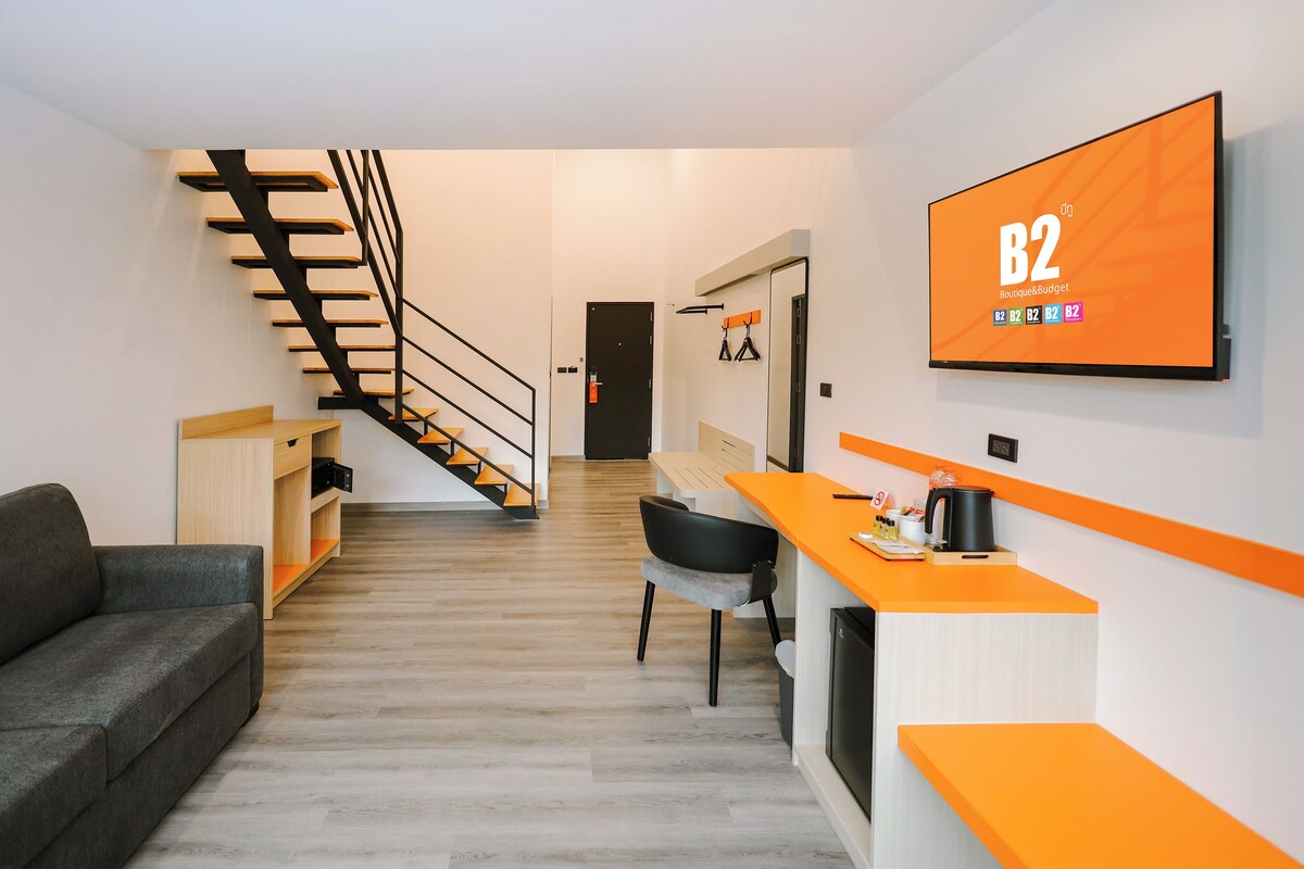 A modern interior with a staircase, a sofa, a desk, and a television displaying 'B2 Boutique & Budget'.