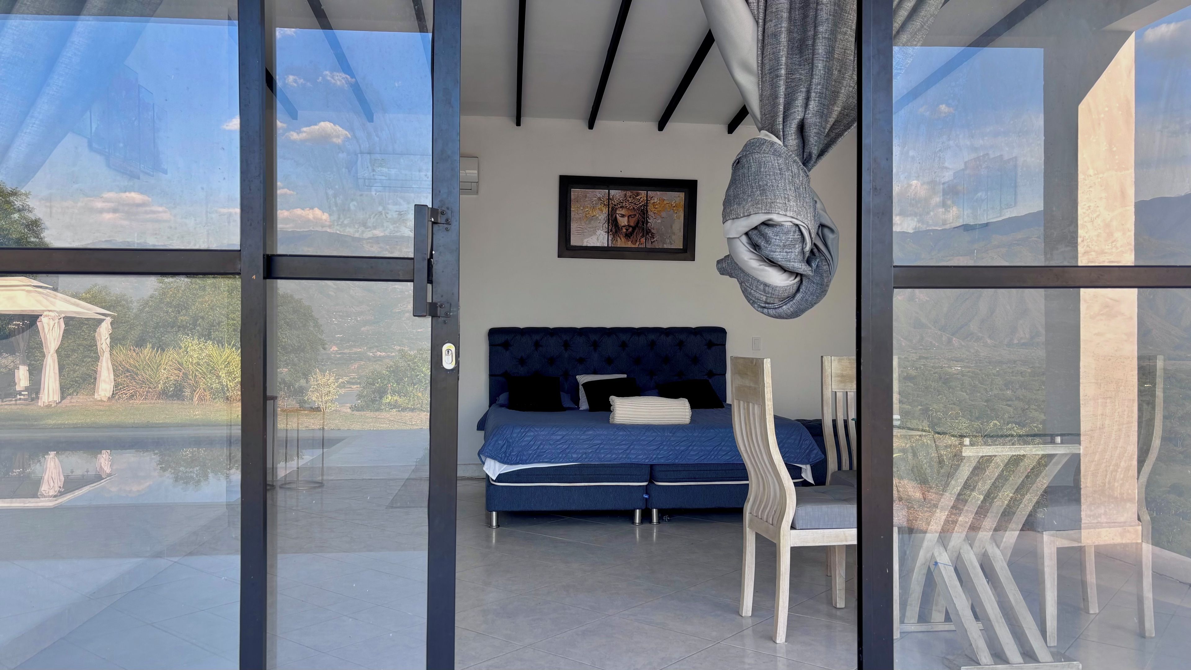 A bedroom with a blue bed, a painting on the wall, and a view through a glass door.