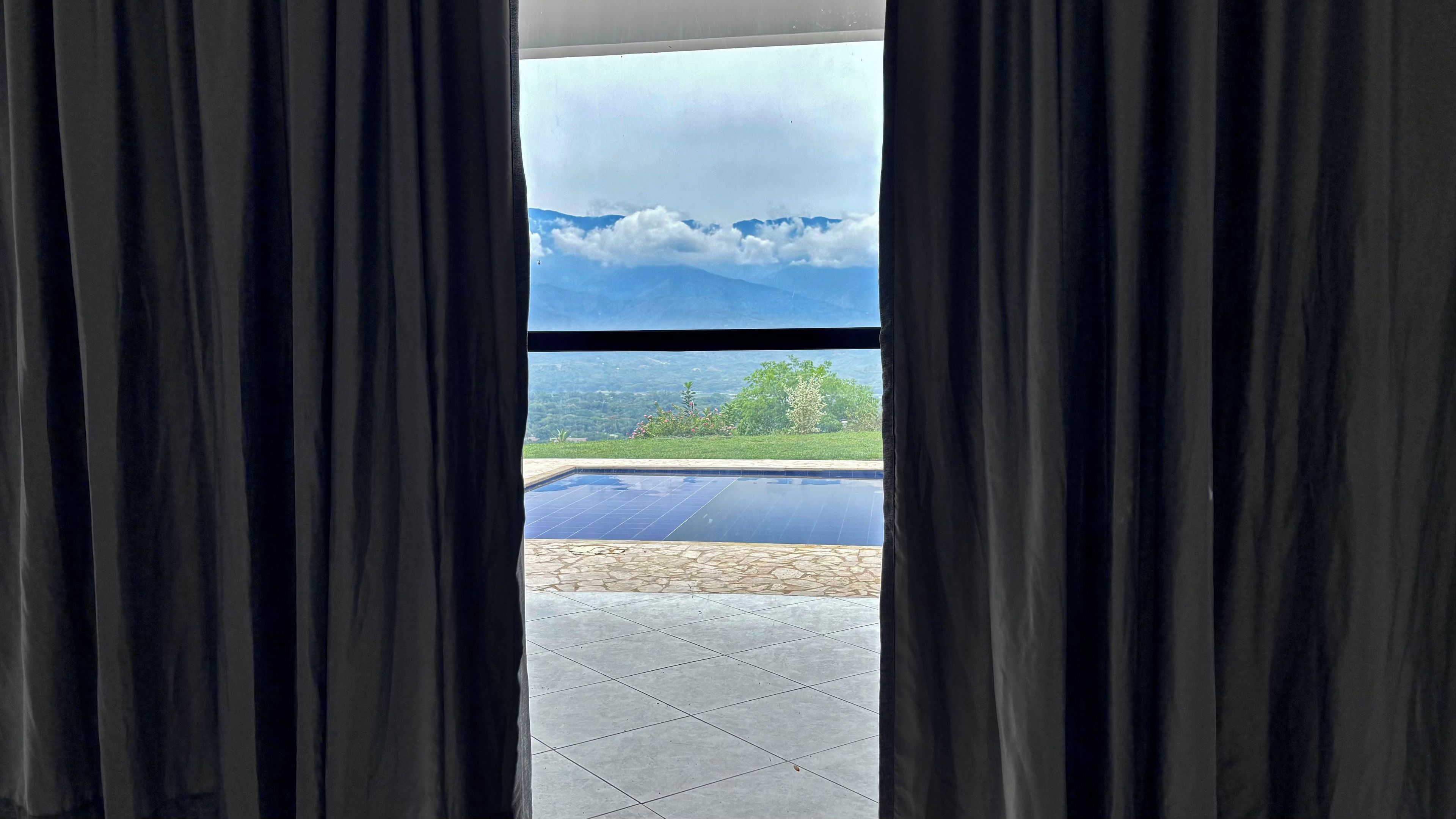 Vista attraverso una finestra con tende, che mostra una piscina e un paesaggio con vegetazione e montagne.