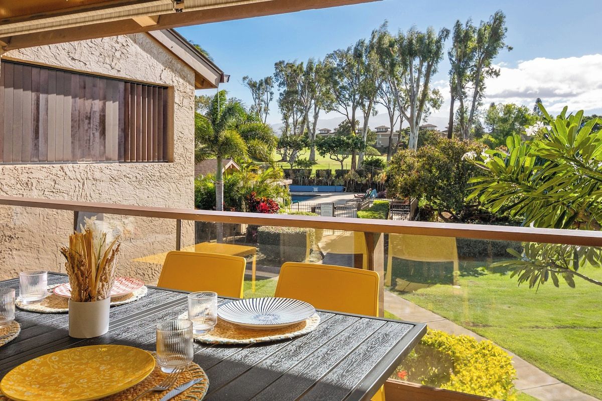 A dining area with a table set for two, overlooking a landscaped garden with palm trees and a swimming pool.