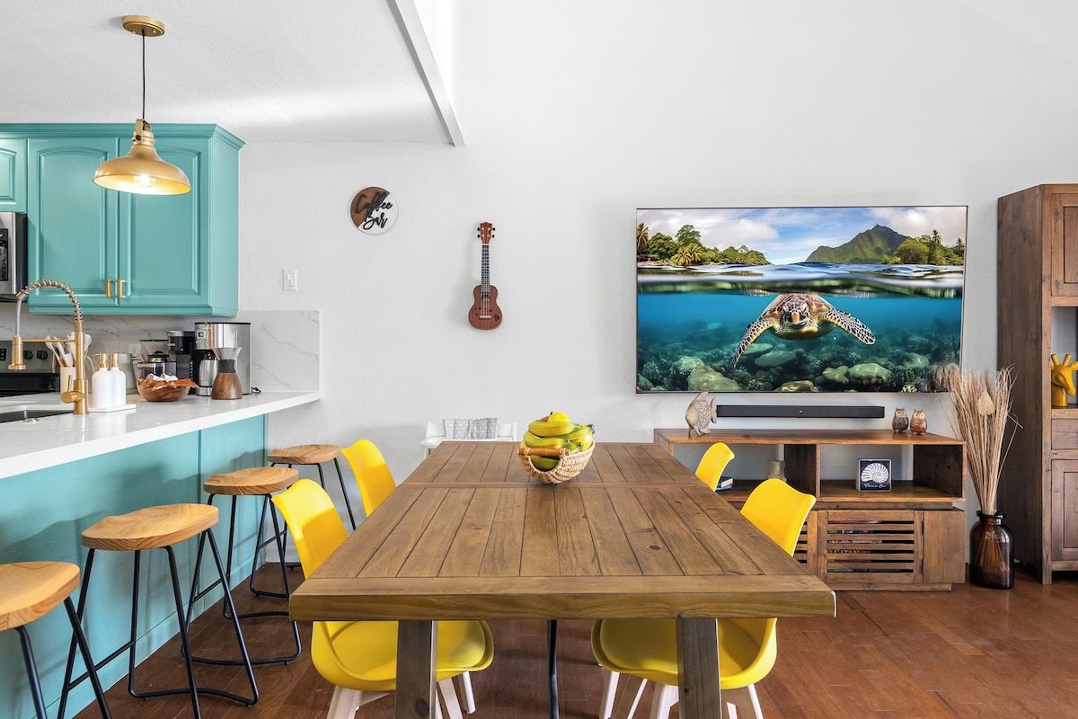 Un coin repas avec une table en bois, des chaises jaunes et un grand téléviseur à écran plat diffusant une scène de plage.