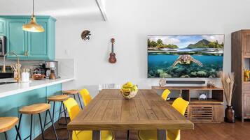 Un coin repas avec une table en bois, des chaises jaunes et un grand téléviseur à écran plat diffusant une scène de plage.