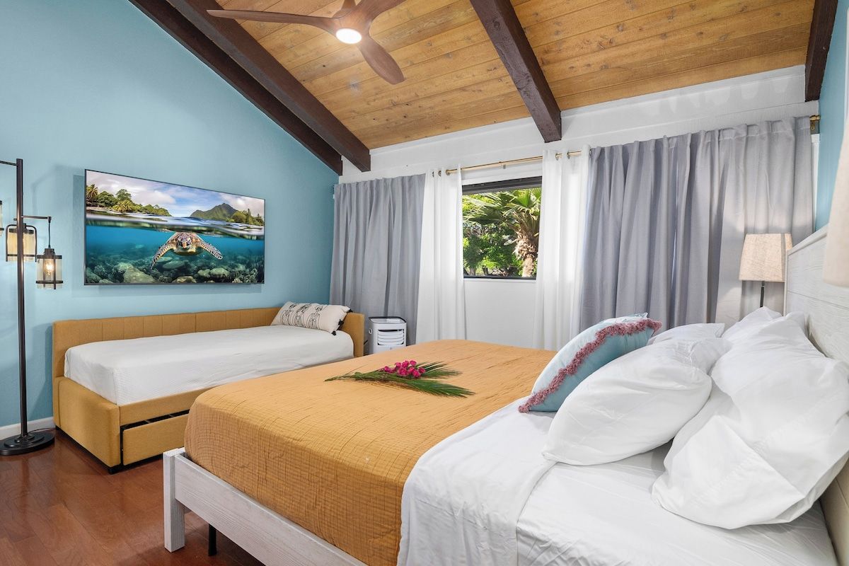 A double bed with a beige and white bedspread, a nightstand with a lamp, a painting of a sea turtle, and a window with curtains.