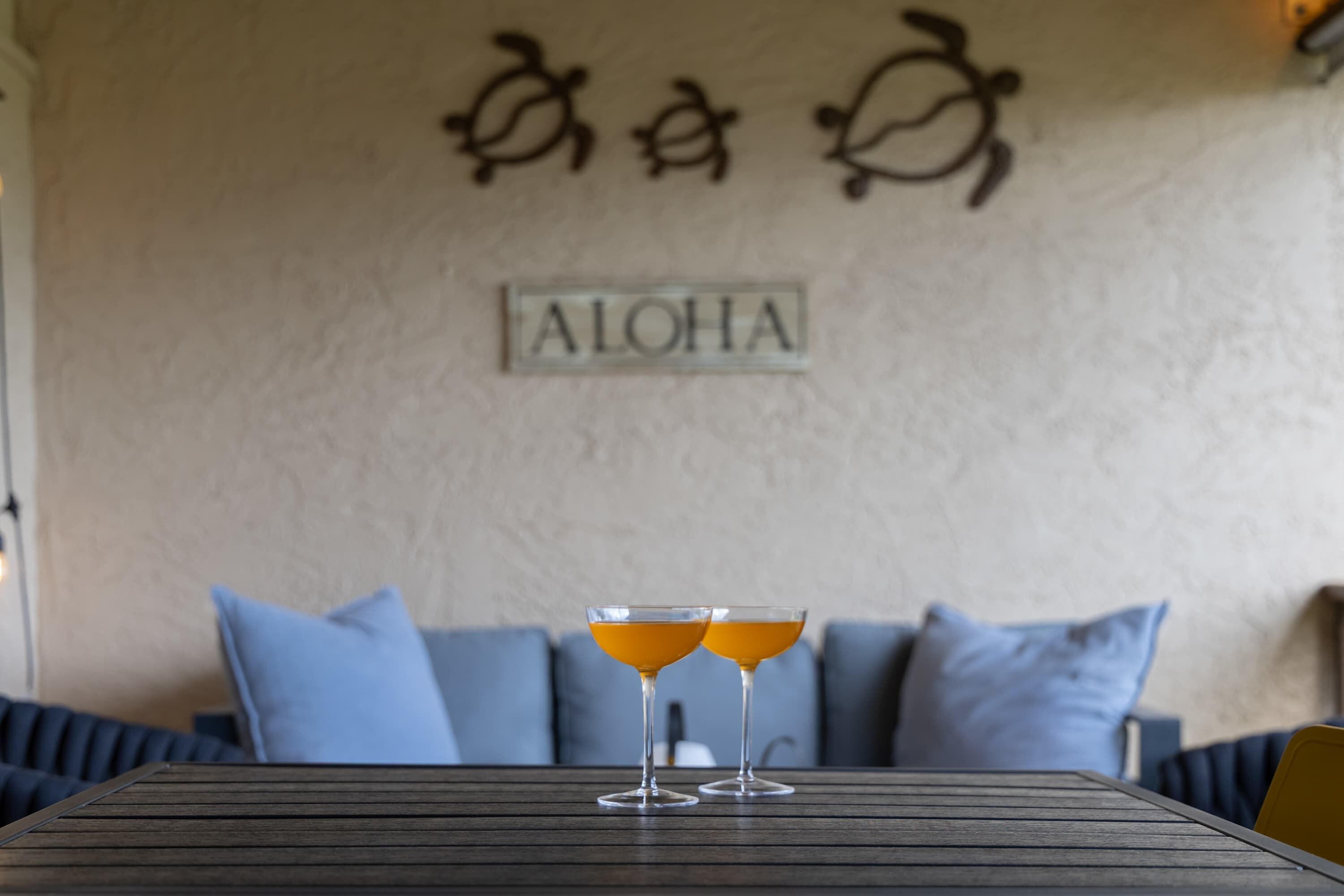 Une table avec deux verres contenant une boisson à l’orange, un mur sur lequel est écrit le mot « ALOHA » et deux motifs décoratifs, et un canapé bleu en arrière-plan.