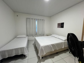 A room with two single beds, a small bedside table, a wall-mounted fan, and a window with curtains. - Cozy, furnished apartment for rent in Porto Seguro. (Porto Seguro)