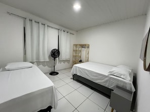 A simple room with two beds, a standing fan, and a window with curtains. - Cozy, furnished apartment for rent in Porto Seguro. (Porto Seguro)