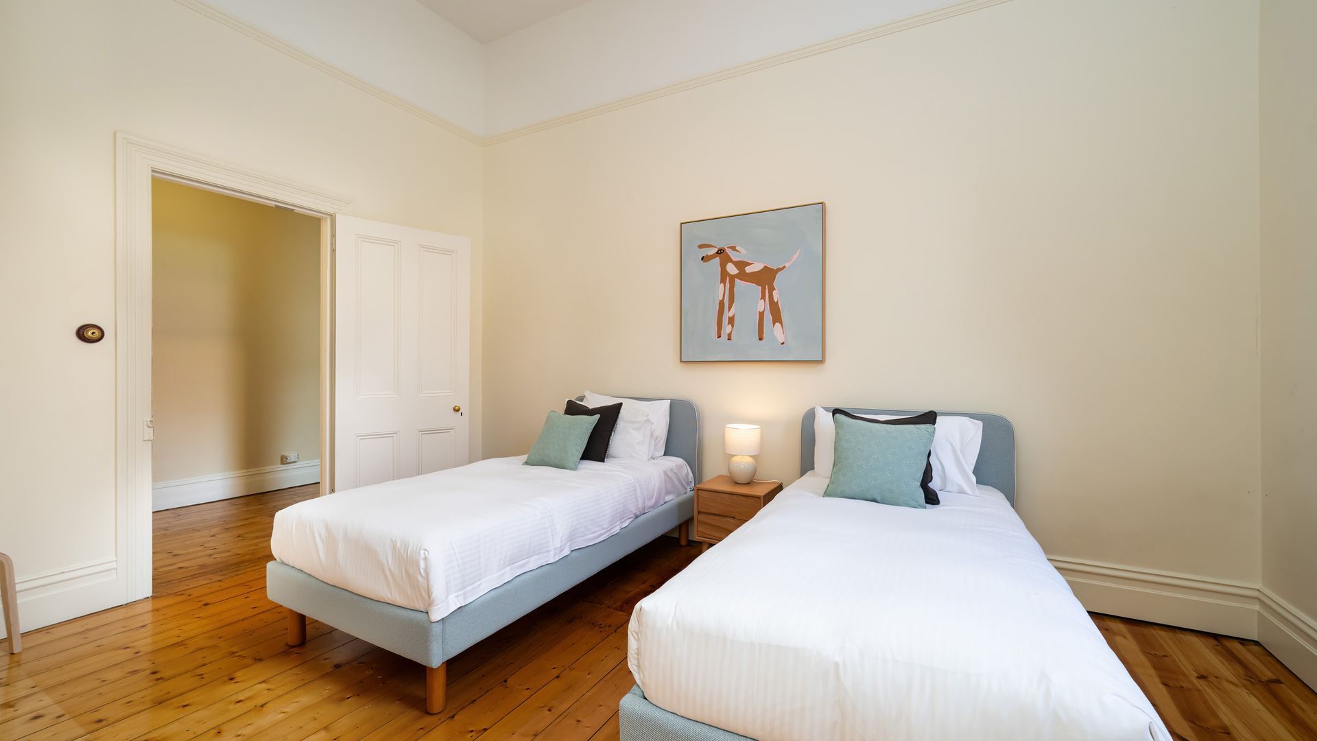 A hotel room with two single beds, a door, a wall-mounted artwork, and a nightstand with a lamp.