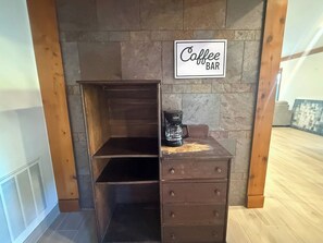 A wooden chest of drawers with a coffee maker on top, a sign that reads 'Coffee BAR', and a stone wall in the background.
