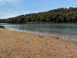 A pebbly beach with a calm lake and a forested hill in the background.