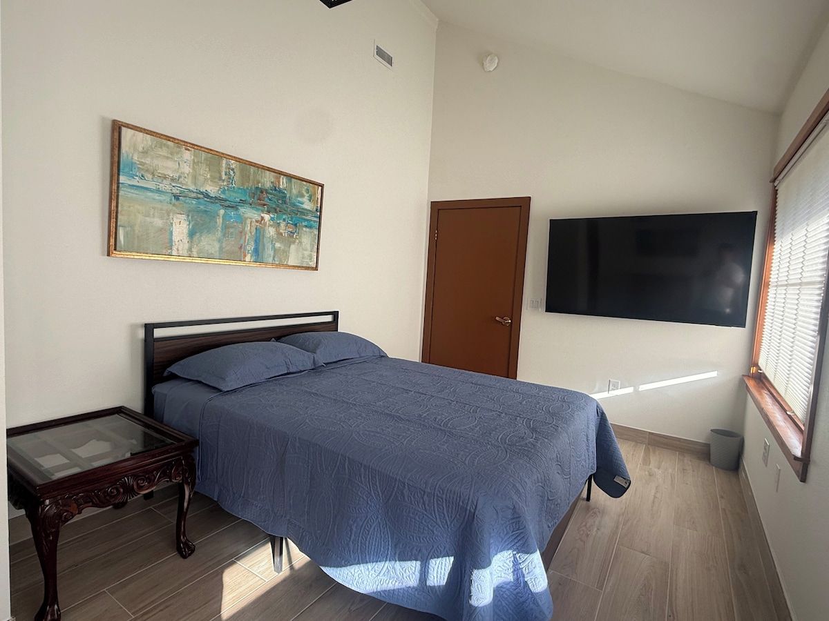 A bedroom with a bed, a bedside table, a painting, a door, and a window.