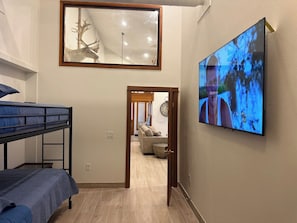 Una habitación con literas, un televisor, un espejo y una puerta que da a una sala de estar con un sofá y un reloj.