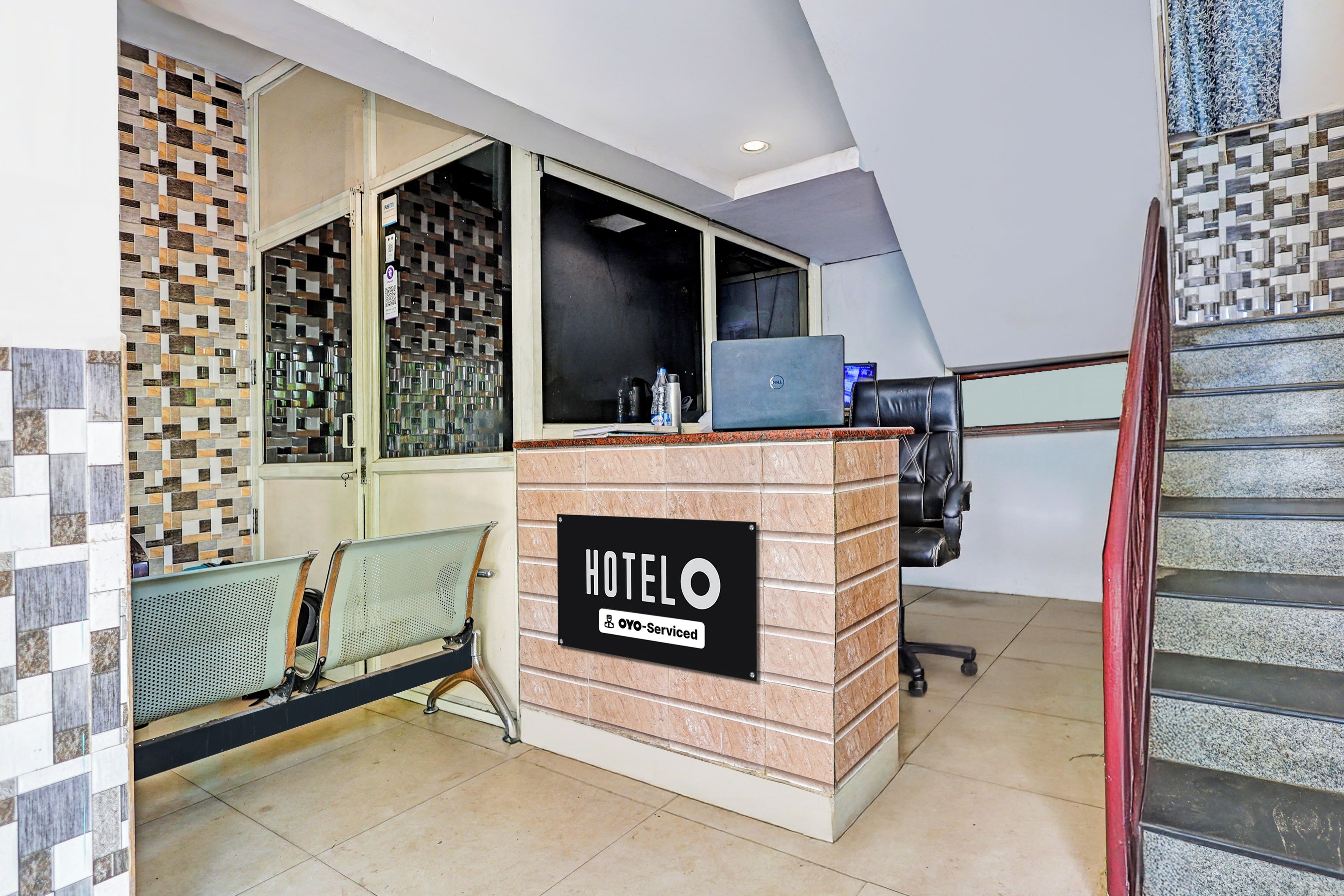 A hotel reception desk with a sign indicating it is serviced by OYO. There is a laptop on the desk, a chair, and a bench with a metal mesh backrest.