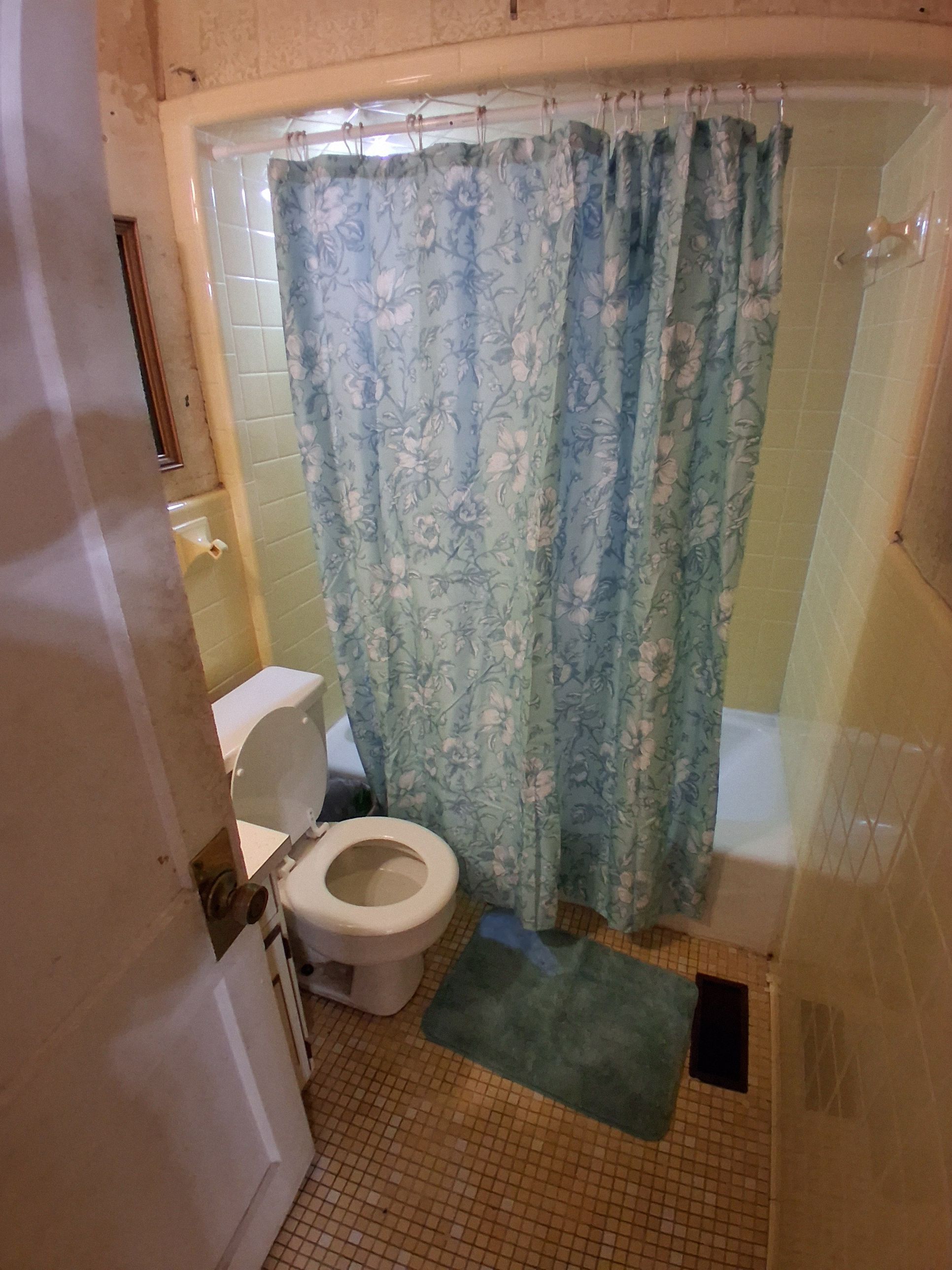 Un baño con inodoro, ducha con cortina estampada y una alfombra pequeña en el suelo.