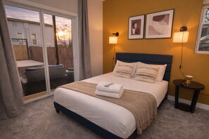 A bedroom with a bed, two wall-mounted lamps, a small table with a lamp, and a sliding glass door leading to an outdoor area.