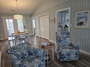 Un salon avec deux fauteuils à motifs, une table à manger avec des chaises, un luminaire suspendu et une vue sur la cuisine par une porte ouverte.