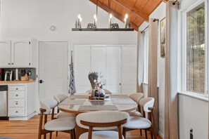 A dining area with a round table, chairs, and a kitchen in the background. - La Cabaña. A Redwood Retreat with Hot Tub & Starry Nights (Pioneer)