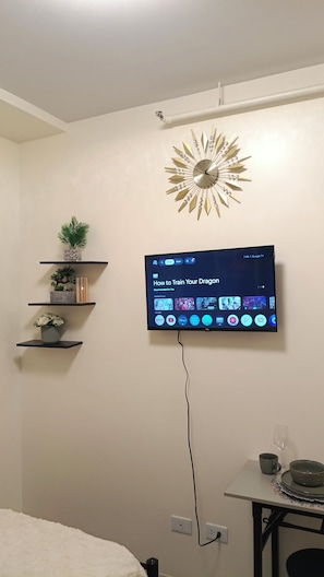 Una stanza con una TV a parete che mostra l'interfaccia di un servizio di streaming, una sveglia decorativa e un piccolo tavolo con un bicchiere.
