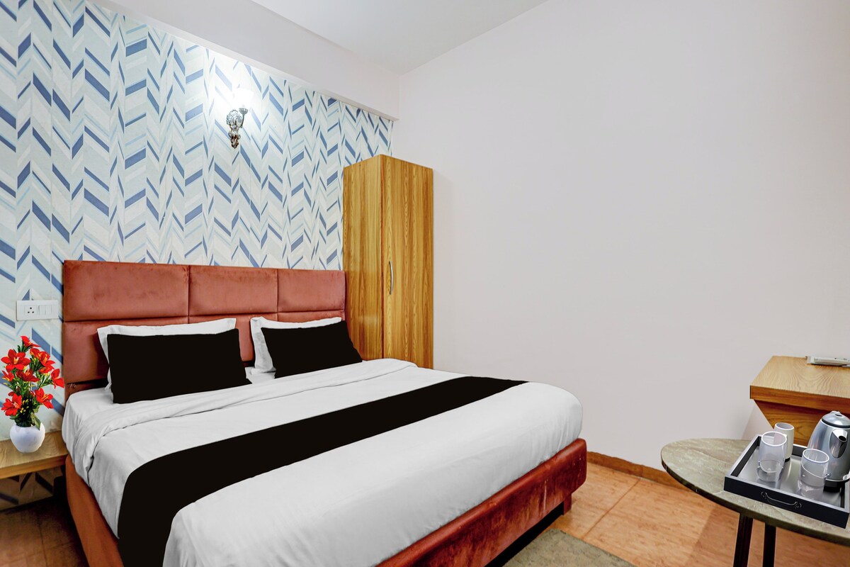 A hotel room with a bed, a wooden wardrobe, a bedside table with a tray, and a wall with a geometric pattern.