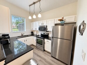 A modern kitchen with white cabinets, stainless steel appliances, a black granite countertop, and a window with blinds. - Regal Oaks Resort | Near Disney & Old Town (Kissimmee)