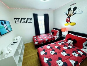 Una habitación con dos camas, cada una con ropa de cama roja decorada con estampados de Mickey Mouse, una pared con un mural grande de Mickey Mouse y un televisor montado en la pared.