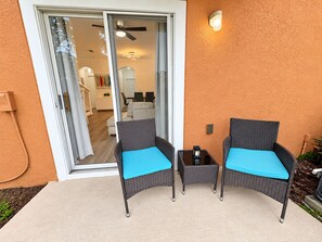 Une terrasse avec deux fauteuils en osier et une petite table, une porte-fenêtre coulissante donnant sur un espace intérieur, et un luminaire fixé au mur.