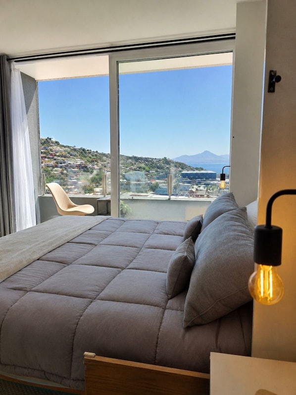 A bedroom with a large bed, a chair, and a view of a hillside and sea.